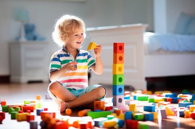 Çocuk renkli oyuncak bloklarıyla oynuyor. Çocuklar oynar. Küçük çocuk güneşli beyaz yatak odasındaki karanlık zeminde oyuncaklardan kule inşa ediyor. Bebek ve bebek için eğitici bir oyun. Çocuklar oyuncak ev yapar.