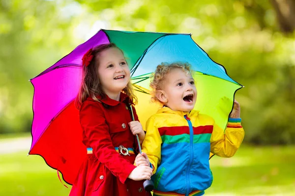 小さな男の子と女の子は雨の夏の公園で遊ぶ 雨の中で遊んでカラフルな虹の傘 防水ジャケットやコートを持つ子供たち 子供たちは秋のシャワーで歩く どんな天気でも屋外の楽しみ — ストック写真