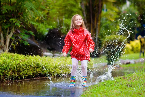 子供は水たまりで遊ぶ 子供たちは秋の雨で屋外で遊んでジャンプします 子供のための秋の雨の天気屋外活動 泥だらけの水たまりに飛び込む子供 防水ジャケットとブーツ用女の子 — ストック写真