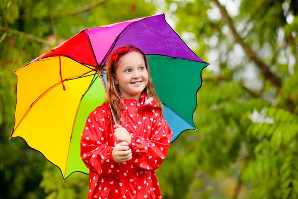 Kid playing in the rain in autumn park. Child with umbrella and rain boots play outdoors in heavy rain. Little girl in red jacket under fall shower. Kids fun by rainy weather. Children play in storm.