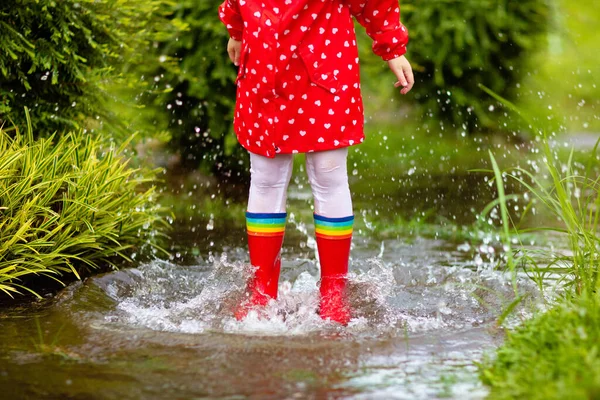 秋の公園で雨の中で遊んでいる子供 子供は雨の日に泥だらけの水たまりに飛び込む 大雨のブーツと赤いジャケットの小さな女の子は屋外で激しいシャワーで 子供防水履物とコート — ストック写真