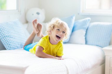 Beyaz güneşli bir odada pencereli bir çocuk yatakta oynuyor. Çocuk odası ve iç tasarım. Erkek bebek evde. Çocuk yuvası için yatak ve tekstil. Oyuncağı ve kitabı olan bir çocuk. Uyku ve uyku zamanı..