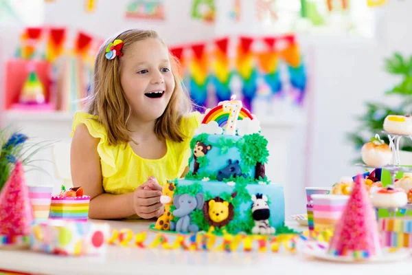 Kids birthday party. Child blowing candles on cake and opening presents on jungle theme celebration. Sweets and cakes for children event. Kid celebrating birthday. Table setting with gifts and pastry.