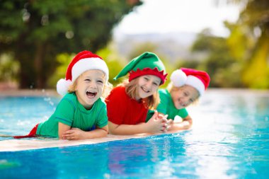 Tropikal adada Noel tatili. Noel Baba şapkalı çocuklar Noel Baba tatilinde yüzme havuzunda oynuyorlar. Kış tatilleri plajda. Çocuklarla seyahat et. Erkek ve kız yüzer. Mutlu Noeller..