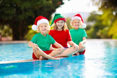 Tropikal adada Noel tatili. Noel Baba şapkalı çocuklar Noel Baba tatilinde yüzme havuzunda oynuyorlar. Kış tatilleri plajda. Çocuklarla seyahat et. Erkek ve kız yüzer. Mutlu Noeller..