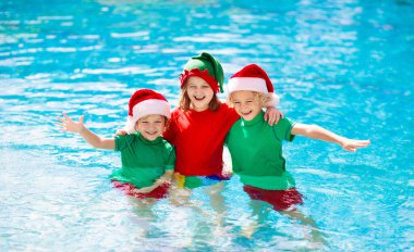 Tropikal adada Noel tatili. Noel Baba şapkalı çocuklar Noel Baba tatilinde yüzme havuzunda oynuyorlar. Kış tatilleri plajda. Çocuklarla seyahat et. Erkek ve kız yüzer. Mutlu Noeller..
