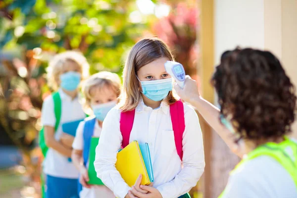 Temperature screening and medical check at school. Child in face mask in class in covid-19 outbreak. Teacher with thermometer at preschool entrance. Social distancing. Coronavirus prevention.