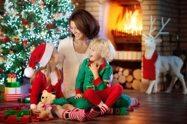 Noel ağacı ve şöminede çocuklu bir aile. Anne ve çocuklar şöminede hediyeleri açıyorlar. Oğlan, kız ve anne hediyeleri açar. Kış tatili iç dekorasyonu. Noel arifesinde pijamalı bir çocuk..