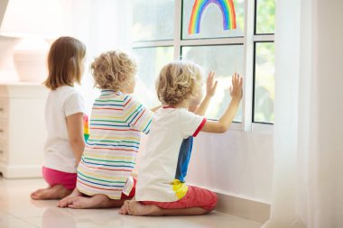 Coronavirus karantinası. Evde kal. Çocuklar pencerede oturuyor. Çocuklar umut gökkuşağı işareti çiziyor. Corona virüsünün tecridi sırasında kız ve erkek. Çocuk ve evcil hayvan. Evde aile izolasyonu. Hastalık önleme