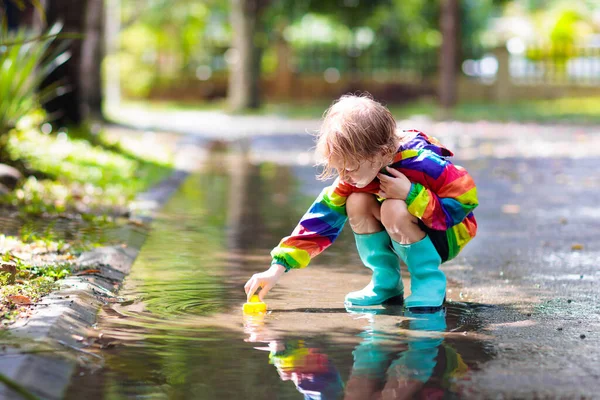 秋の公園で雨の中で遊んでいる子供 子供は雨の日に泥だらけの水たまりに飛び込む 大雨のブーツと赤いジャケットの小さな男の子が屋外で激しいシャワーで 子供防水履物とコート — ストック写真