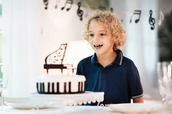 Music and piano theme kids birthday party. Kid with black and white cake. Child blowing candles and opening present. Little boy celebrating birthday. Event for young pianist. Festive decoration.