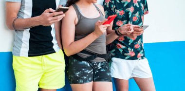 Üç genç, yaz kıyafetleri içinde, beyaz ve mavi bir duvarda, akıllı telefonlarını kullanıyorlar.