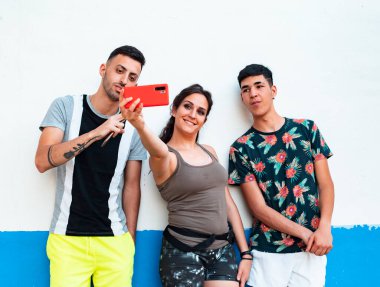 Üç genç, yaz kıyafetleri içinde, beyaz ve mavi bir duvarda, akıllı telefonlarını kullanıyorlar.