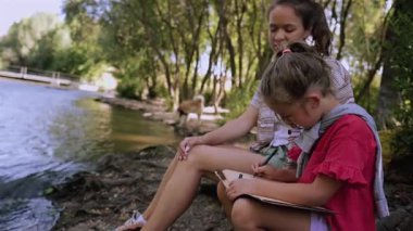 Older sister guiding younger sibling while drawing together beside flowing river, surrounded by lush natural landscape during peaceful summer day