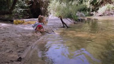 Young blonde girl playing joyfully with small white dog on sandy riverbank near shallow creek, surrounded by sunlit forest trees with clear water flowing nearby