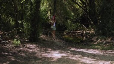 Sunlight filtering through forest canopy, mother carrying son on shoulders while walking along tranquil woodland path beside gentle stream, sharing playful outdoor adventure