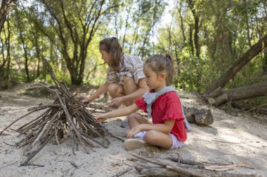 Mother and daughter building a campfire together in a serene forest, sharing moments of teamwork and creating cherished memories while enjoying the beauty of nature