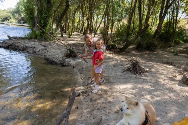 Grandmother teaching her granddaughter how to fish on the shore of a river with their akita dog sitting nearby, enjoying a sunny day in nature