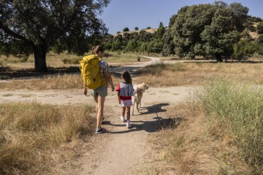 Mother with yellow backpack and daughter walking with their dog on a dirt road in the countryside, enjoying a summer hike surrounded by dry grass and trees