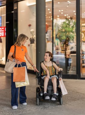 Two women are walking and talking in a shopping mall while carrying shopping bags, one of them is using a wheelchair and they are both smiling and looking at each other