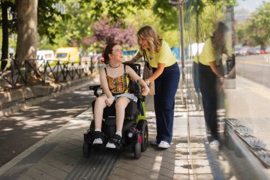 Happy volunteer assisting a smiling young disabled woman on a wheelchair while waiting at a bus stop, enjoying a friendly conversation on a sunny summer day