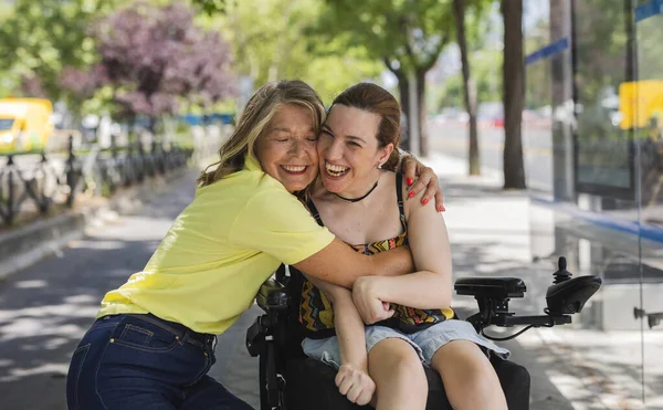 Senior woman volunteer is embracing and sharing a tender moment with a happy disabled young woman in a wheelchair, enjoying a sunny day outdoors near a bus stop