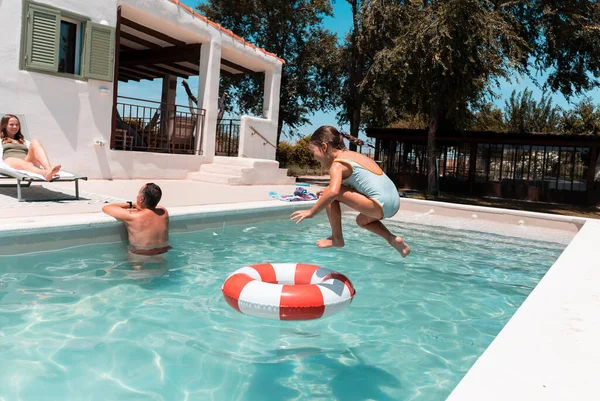 Girl joyfully jumping into a swimming pool while her father and mother relax by the poolside, enjoying a sunny summer vacation day filled with laughter and family fun