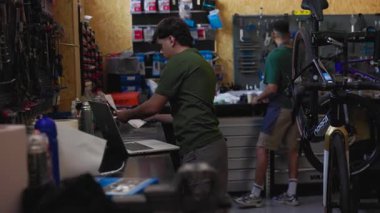 Young male mechanic processing online orders using laptop while colleague performs bicycle maintenance in bustling workshop. Demonstrating collaborative small business efficiency and professionalism