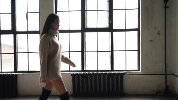 Steadicam lövés egy fiatal gyönyörű ázsiai lány egy nagy hangulat a munkába való út. Modell ázsiai megjelenése, egy hosszú pulóvert sétál az ablak előtt egy gyönyörű Loft. Lassított mozgás. — Stock videók