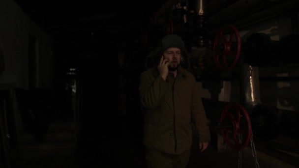 Russian man in a hat with earflaps, talking on the phone in a dark basement and decides matters of national importance. General plan — Stock Video