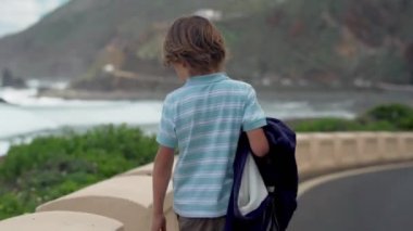Kırsal yolda yalnız çocuk yürüyüşleri arka görünümü. Çocuk yürüyüş ve dağlar ve deniz manzarası bakıyor.