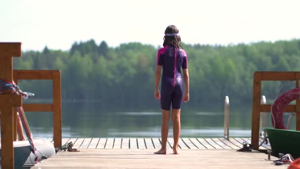 Chica en traje de neopreno saltando al lago desde el muelle de madera. Divertirse en el día de verano — Vídeo de stock