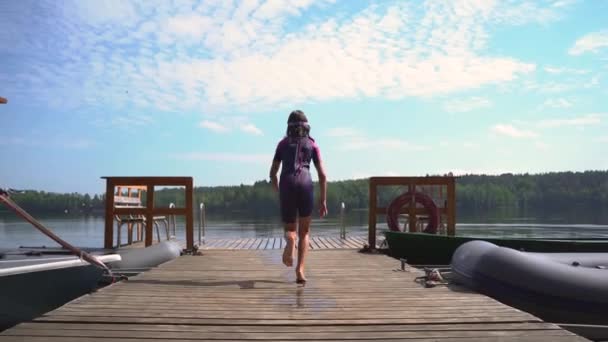 Dívka v neoprénu skočí z dřevěného mola do jezera. Bavíte se v letní den — Stock video