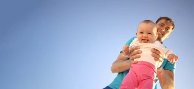 Babam küçük bir çocuğu kollarında tutuyor. Yazın mavi gökyüzünün arka planında. Sevimli bebek gülümsüyor. Dışarıda mutlu bir aile.