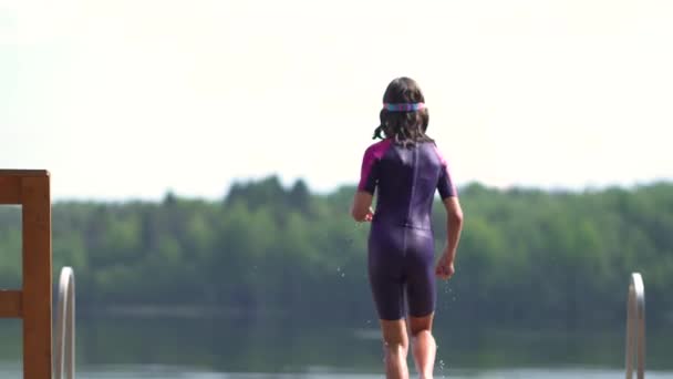 Chica en traje de neopreno saltando al lago desde el muelle de madera. Divertirse en el día de verano — Vídeo de stock