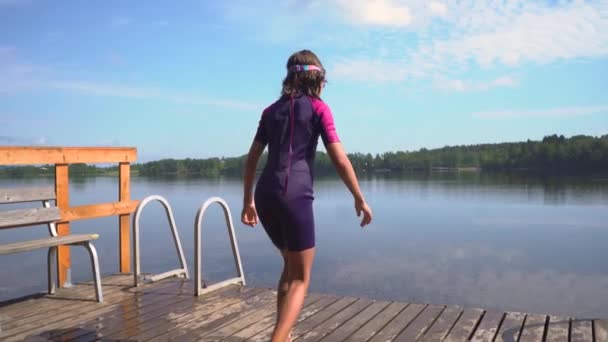 Chica en traje de neopreno saltando al lago desde el muelle de madera. Divertirse en el día de verano — Vídeos de Stock