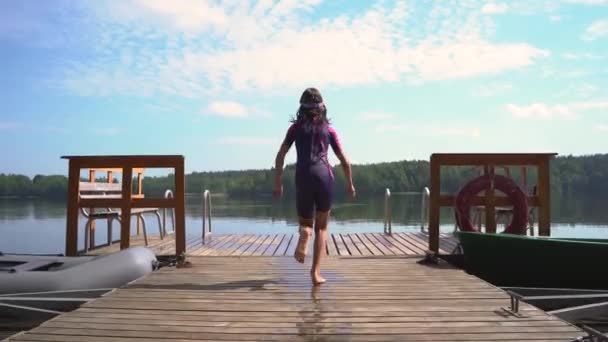 Девушка в гидрокостюме прыгает в озеро с деревянного пирса. Веселитесь в летний день — стоковое видео