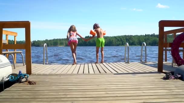 Дівчинка і маленький хлопчик в гідрокостюмі стрибають в озеро з дерев'яного пірсу. Розважаючись в літній день . — стокове відео