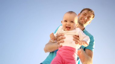 Babam küçük bir çocuğu kollarında tutuyor. Yazın mavi gökyüzünün arka planında. Sevimli bebek gülümsüyor. Dışarıda mutlu bir aile.