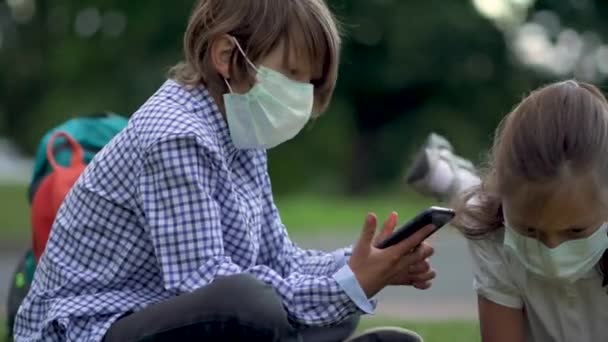 Roztomilí žáci používají chytré telefony na základní škole. Chlapec a dívka v bezpečnostních maskách s pomůckami v rukou. — Stock video