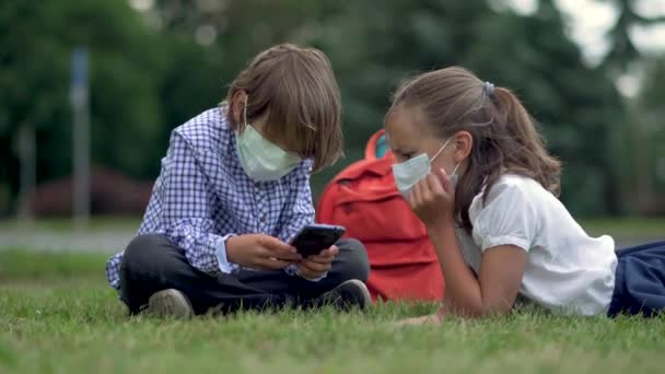 Aranyos diákok okostelefonokat használnak az általános iskolában. Fiú és lány biztonsági maszkban, kütyükkel a kezükben.. — Stock videók