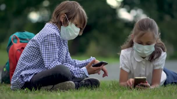 Aranyos diákok okostelefonokat használnak az általános iskolában. Fiú és lány biztonsági maszkban, kütyükkel a kezükben.. — Stock videók