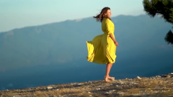 Bella giovane donna in un abito giallo è in piedi su una scogliera di una montagna. Il vestito e i capelli svolazzano nel vento. — Video Stock