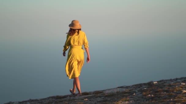 Bella giovane donna in un abito giallo è in piedi su una scogliera di una montagna. Il vestito e i capelli svolazzano nel vento. — Video Stock