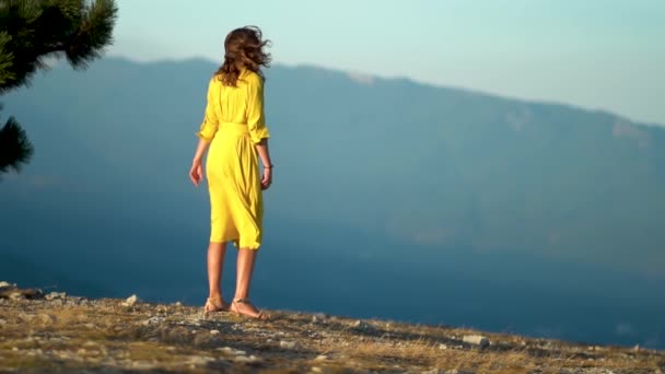 Vacker ung kvinna i gul klänning står på en klippa av ett berg. Klänningen och håret fladdrar i vinden. — Stockvideo