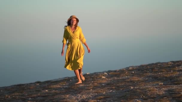 Bella giovane donna in un abito giallo è in piedi su una scogliera di una montagna. Il vestito e i capelli svolazzano nel vento. — Video Stock
