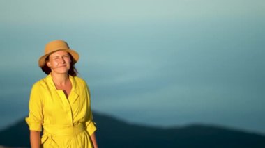 Sarı elbiseli güzel genç bir kadın bir dağın yamacında duruyor. Elbise ve saçlar rüzgarda savruluyor.