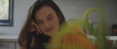 Üzücü bir şekilde gülümseyen, üzgün ve incinmiş genç bir kadın evdeki mutfak masasında otururken. Ayrılmak, aşk, nefret, boşanma kavramı. Yaklaş, Sığ DOF, Yavaş çekim, BMPCC 4K