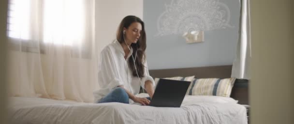 一位年轻女子一边在笔记本电脑上工作一边用耳机与人交谈 面带微笑 从家里工作的概念 慢动作 Bmpcc — 图库视频影像