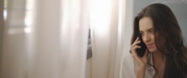 Beyaz gömlekli bir kadının telefonda biriyle konuşurken, gülümserken, pencerenin yanında bir yatakta otururken. Ağır çekim, BMPCC 4K